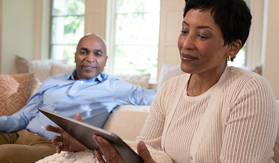 A woman reads on a tablet while a man smiles on the chair next to her.