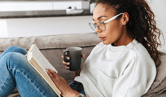 A woman reads while drinking coffee on a couch.