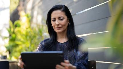 A woman reads on a tablet while sitting at an outdoor table.