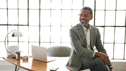 A smiling man sits cross legged on top of desk.