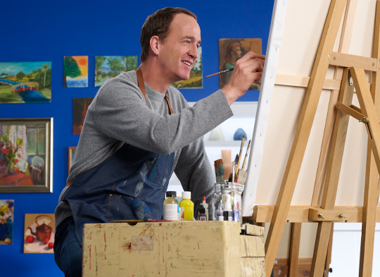 Peyton Manning wears a smock and holds a paintbrush while happily painting in an art studio.