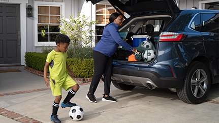 A person in a blue shirt loading a soccer ball into the trunk of a car, while a child in a yellow jersey stands nearby with another soccer ball