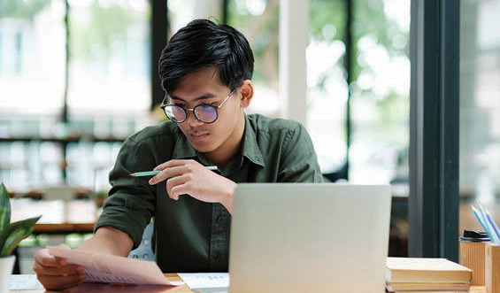 Man with glasses sits at computer, looking at a file. 