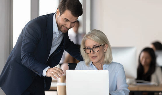 A man points to a computer while speaking to a woman in an office.