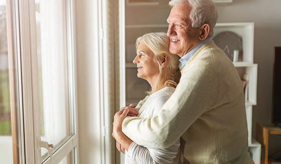 An elderly couple embraces as they look out of a window.