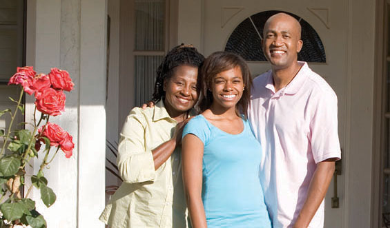 A Gen X couple stands on their front porch with their teenage daughter, smiling.