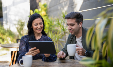 Man and woman sitting outdoors. The woman is holding a tablet and showing the man something on the screen. He's holding a coffee mug.