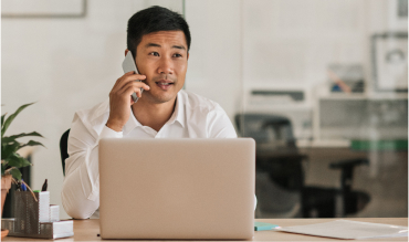 Man is sitting at a desk behind a laptop. He is on a cell phone.